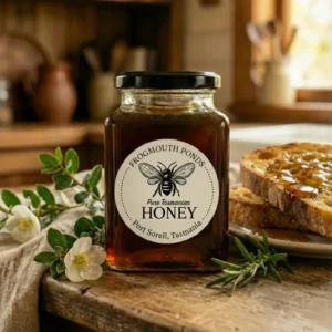 Jar of Frogmouth Ponds Pure Tasmanian Honey (dark, rich Leatherwood variety) next to fresh white flowers and rosemary, with a slice of toast topped with honey in the background, set in a country kitchen.